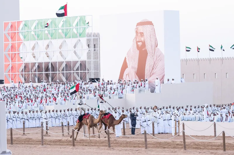 Emirati riders performing camel stunts on parade track in front of grandstand and Sheikh Zayed portrait wall