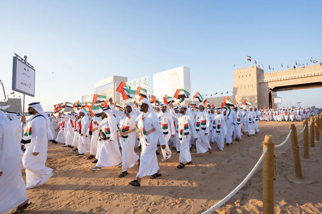Horsemen carrying UAE national flags riding along sand parade track in front of grand fortress walls