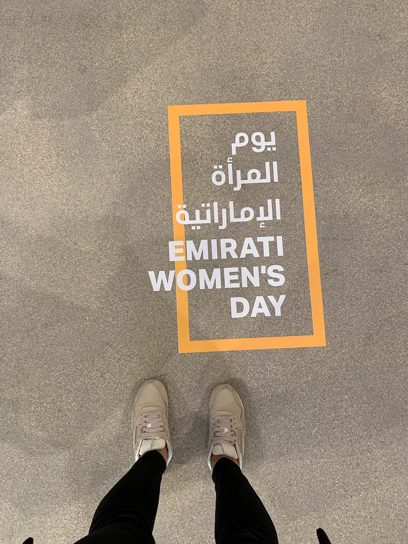 Emirati Women’s Day bilingual floor graphic framed in orange, viewed from above with a visitor standing nearby