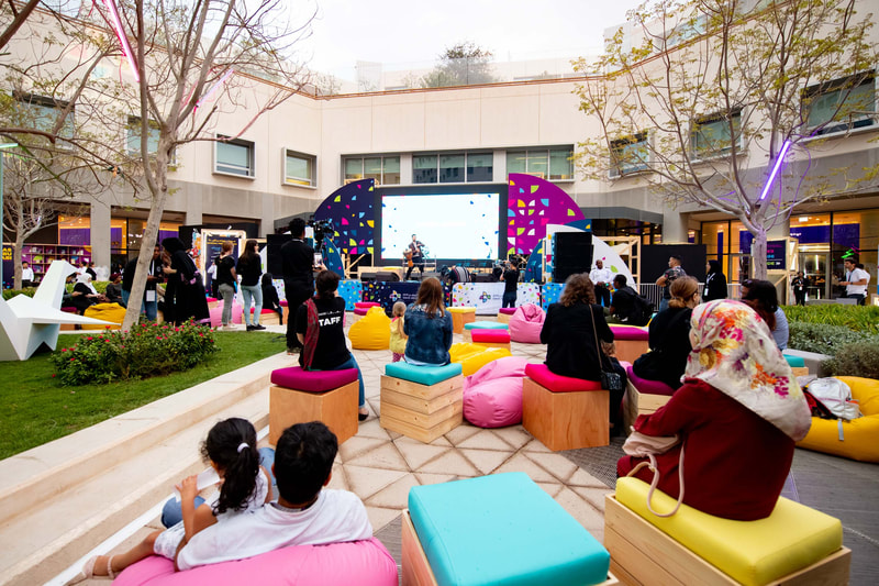 Image of a crowd of people sitting in a open air forum facing an entertainer on the stage. Bright decor.