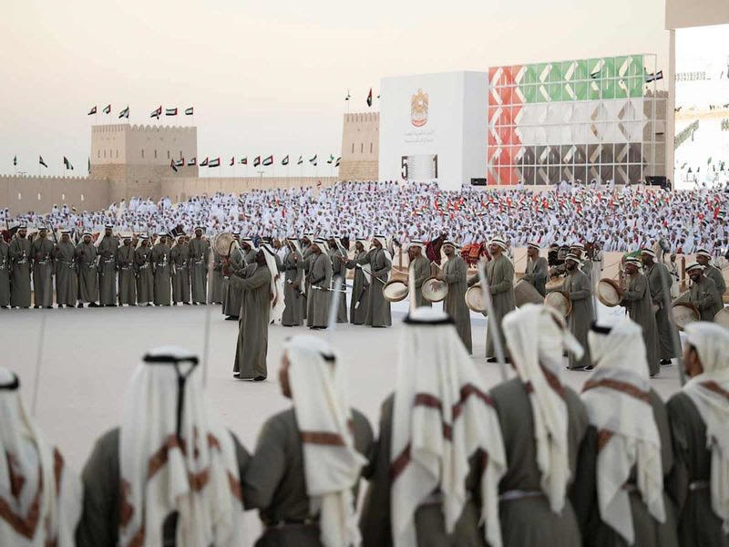 Crowd of Emiratis holding sabers with a large backdrop of the UAE flag